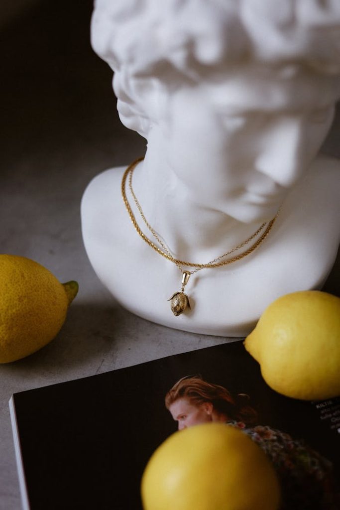 A marble bust adorned with jewelry among lemons and a magazine.